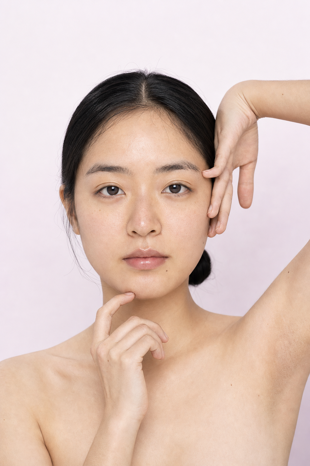 Woman with her hair pulled back posing with one arm raised against a soft pink background